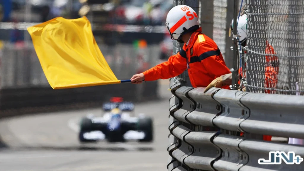 F1黃旗事故警示實景，Marshal工作人員在賽道彎道護欄後方揮舞黃色旗幟，背景有F1賽車減速通過危險路段。